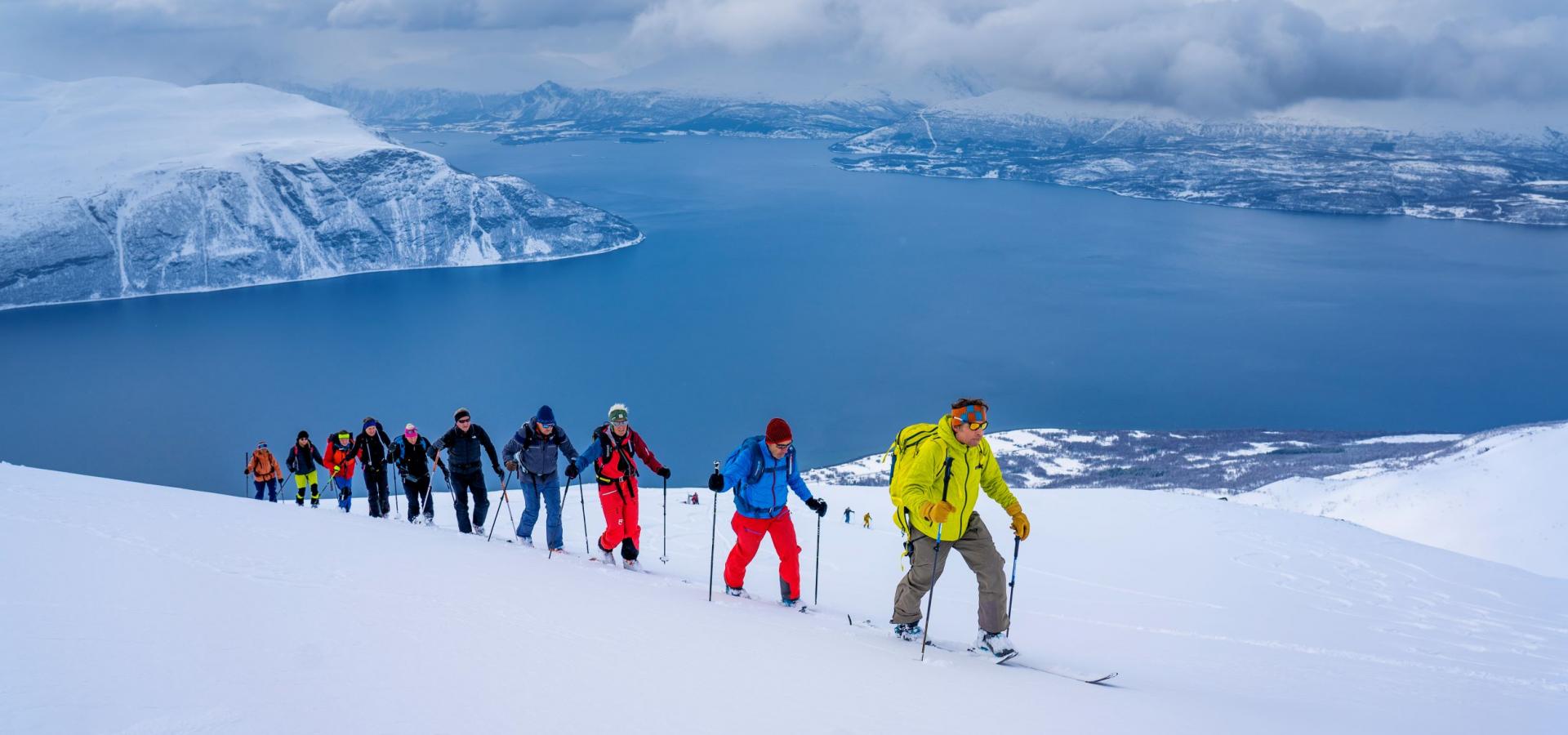 10 persons skiing. Walking up the mountain. Sea behind them.