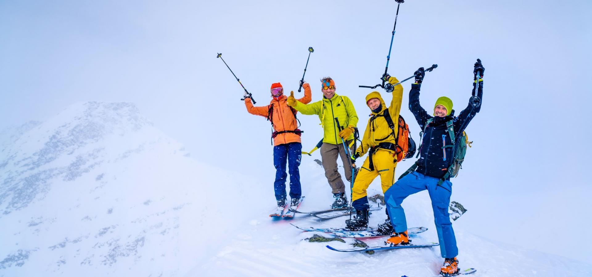 Four people on a skiing trip, standing on the top of a snowy mountain. They are all wearing bright coloured clothes. They look very happy. Some of them have their arms up in the air. 