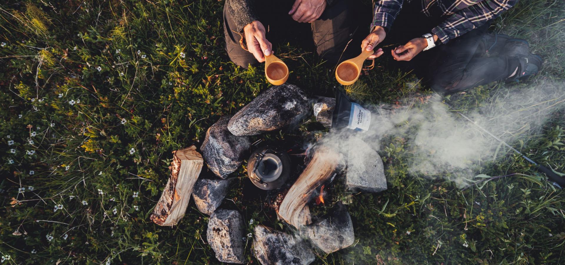 Two hands holding coffee mugs by a campfire