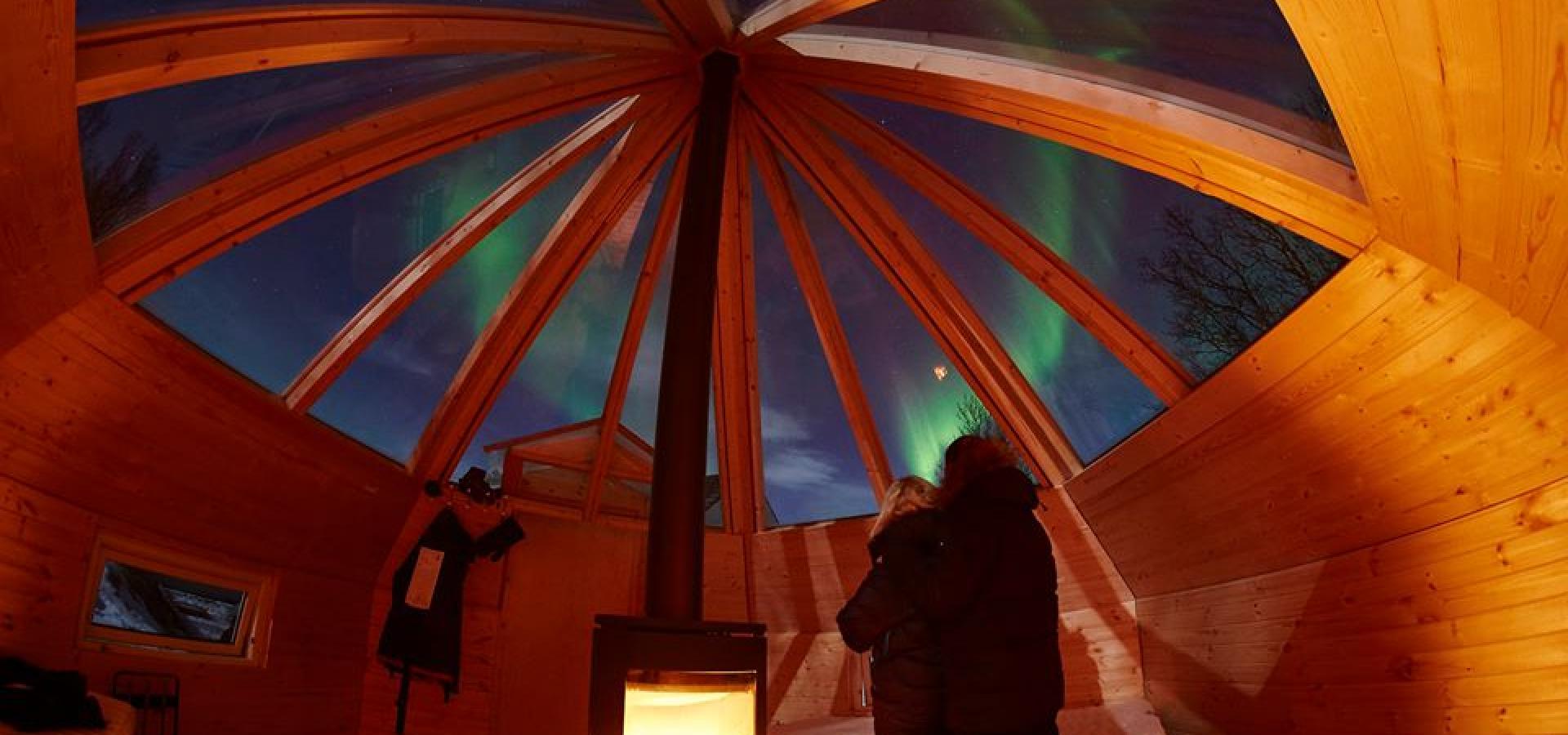 Two persons embrazing and looking up at the northern lights, through the glass roof of the wooden iglo