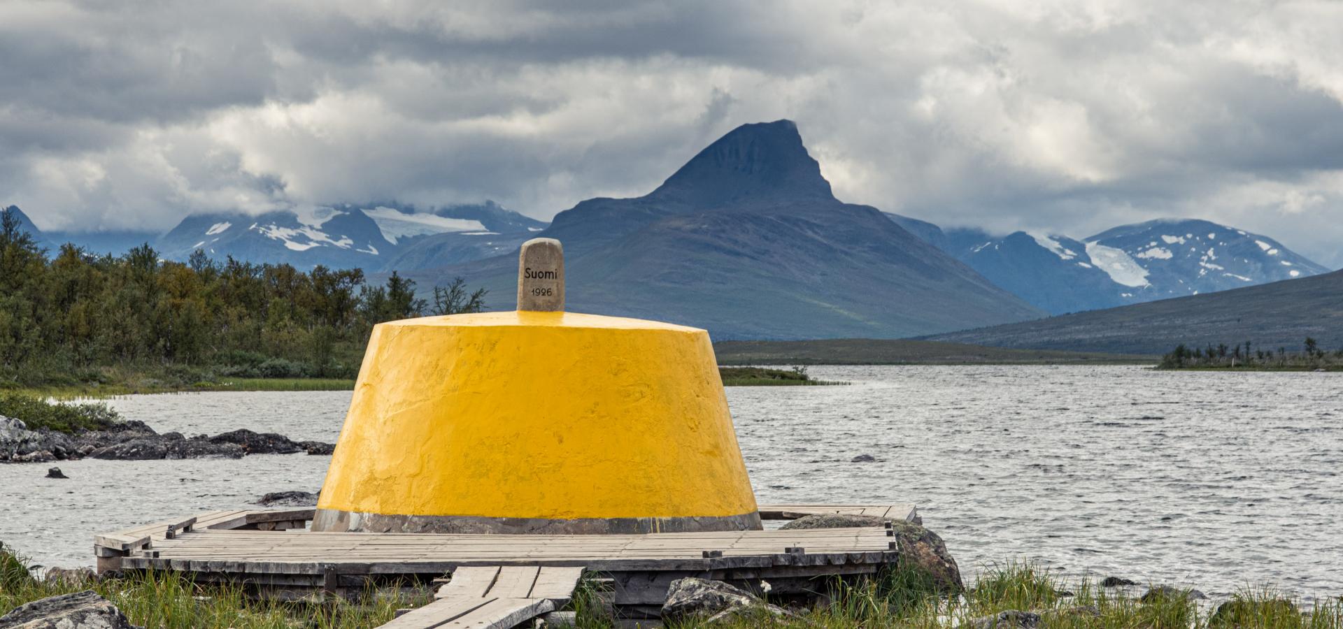 The yellow painted border marker at the three country cairn with the lake and norwegian mountains in the background