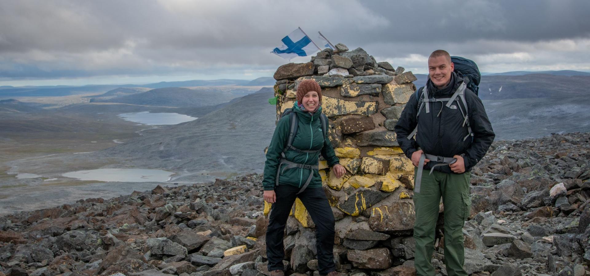 Two hikers by the cairn at Halti, with a view towards some lakes