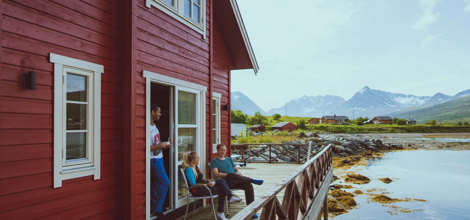 A red cabin by the fjord and 3 persons enjoying life outside on the pier, mountains in the background