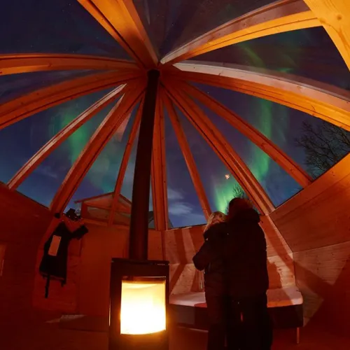 Two persons embrazing and looking up at the northern lights, through the glass roof of the wooden iglo