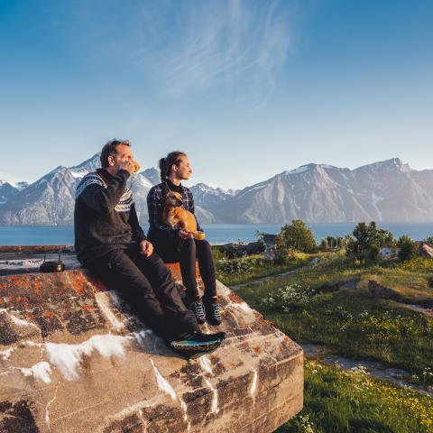 Two people sitting on a big piece of concrete. Its summer, and you can see the lyngen alps. There is also a little dog on the womans lap.