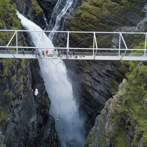 Long metal bridge over a deep canyon and a waterfall on the background.