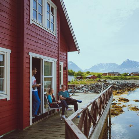 A red cabin by the fjord and 3 persons enjoying life outside on the pier, mountains in the background