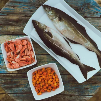 Shrimps in a bowl, cloudberry in a bowl and two fish on a plate placed on a wooden table, picture taken from above