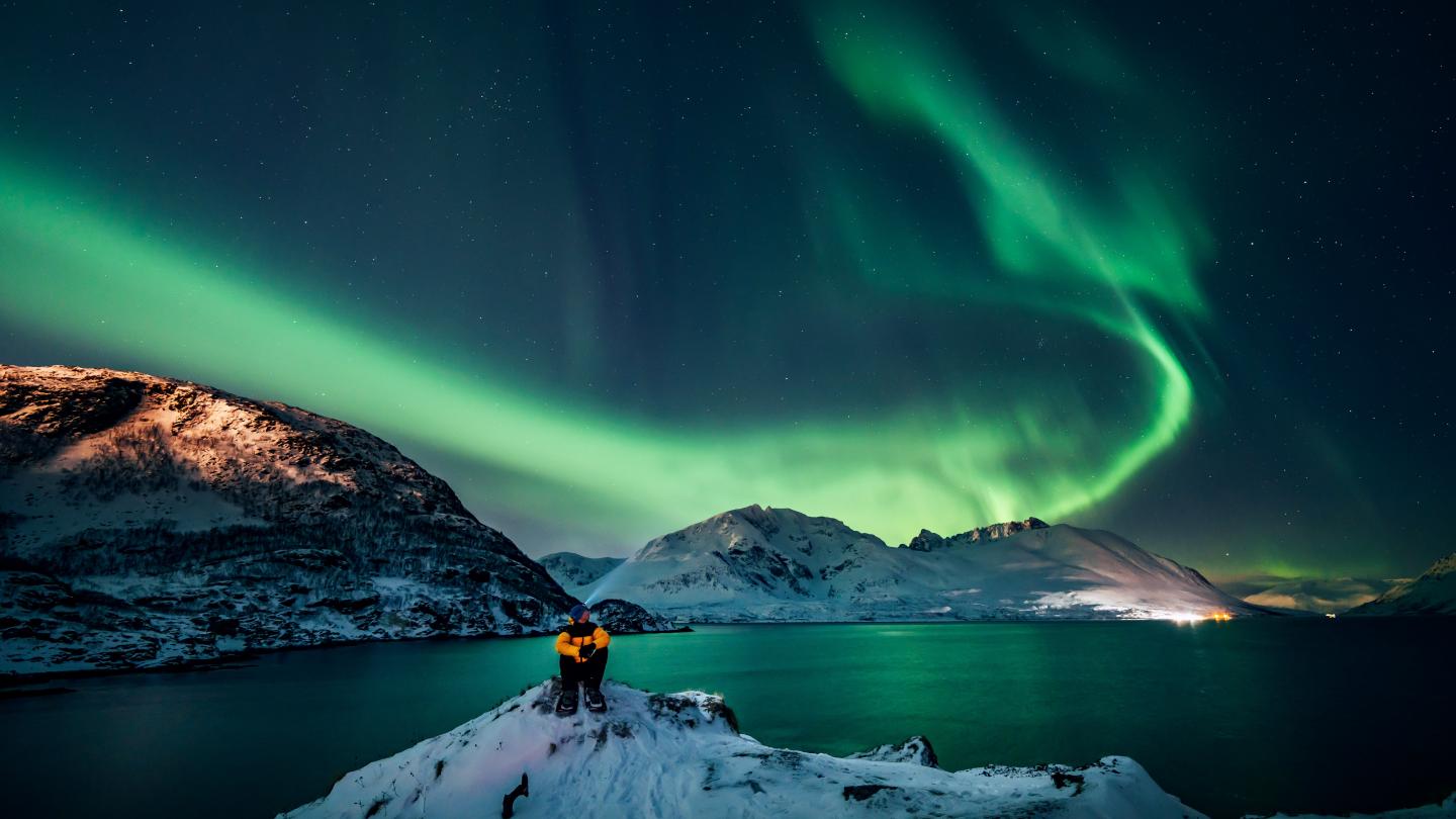 Man sitting on a big rock, behind him you can see mountains and northern lights