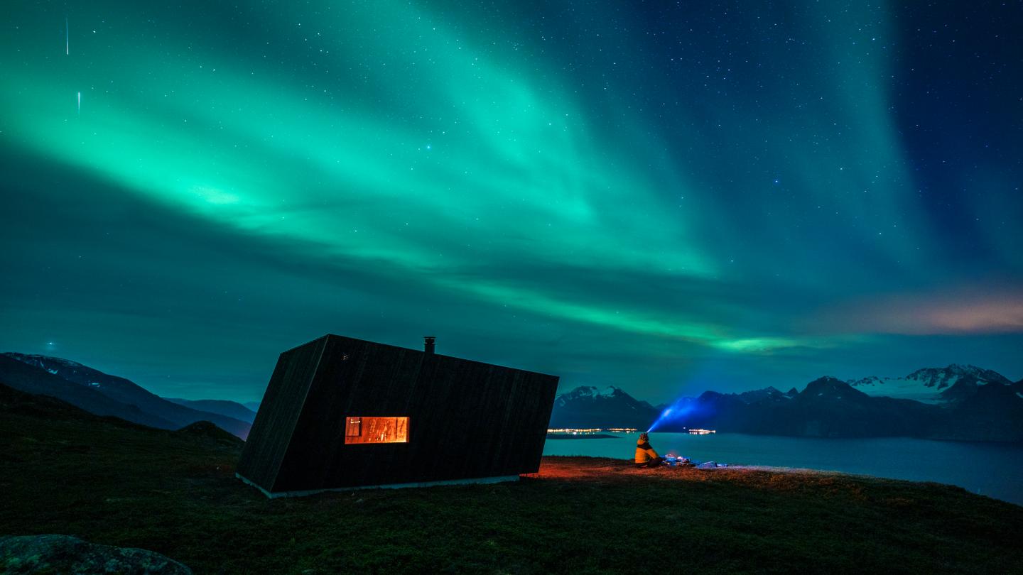A man standing by a cabin watching the northernlights dancing over the Lyngenfjord and the Lyngen Alps in the background