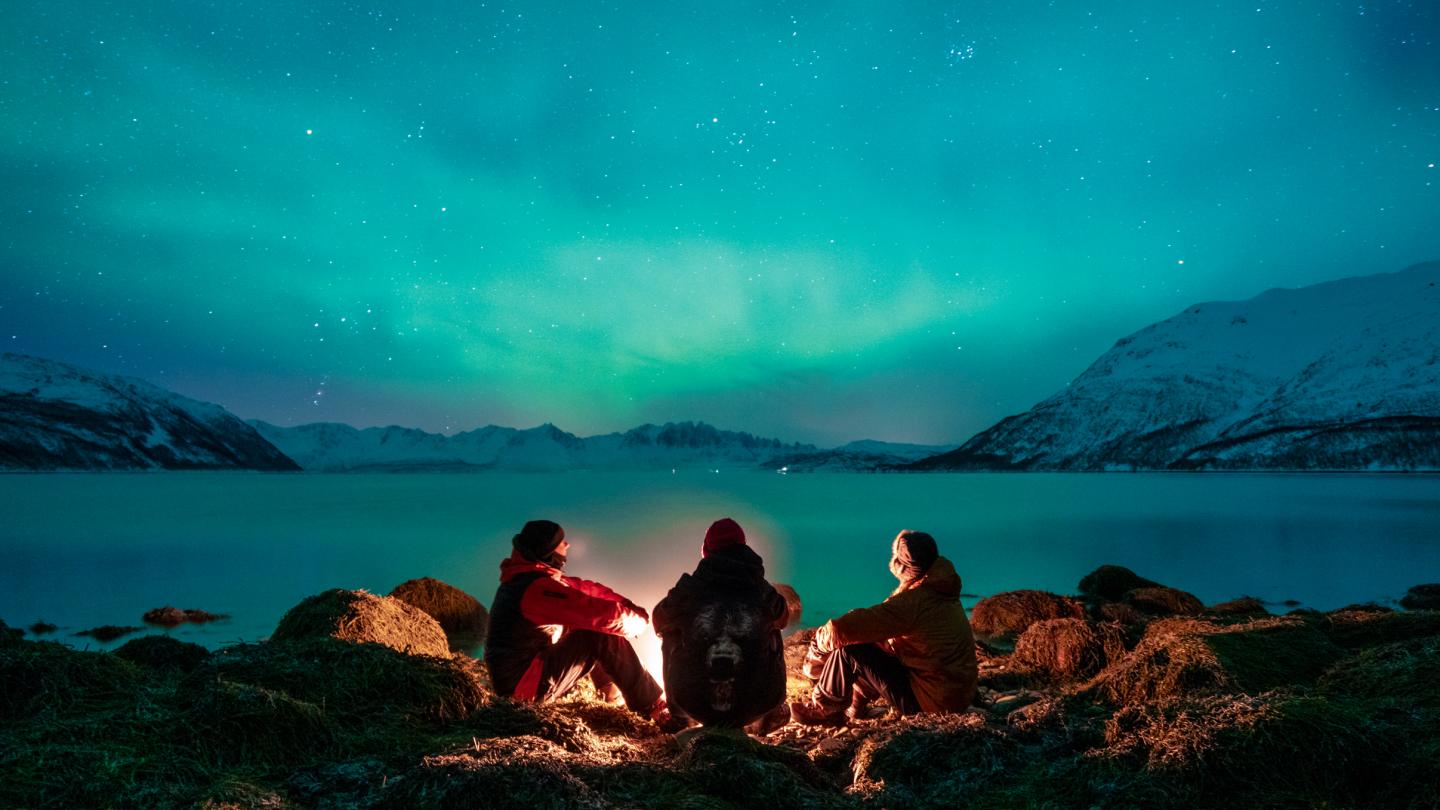 People sitting around camp fire and green northern lights on the sky