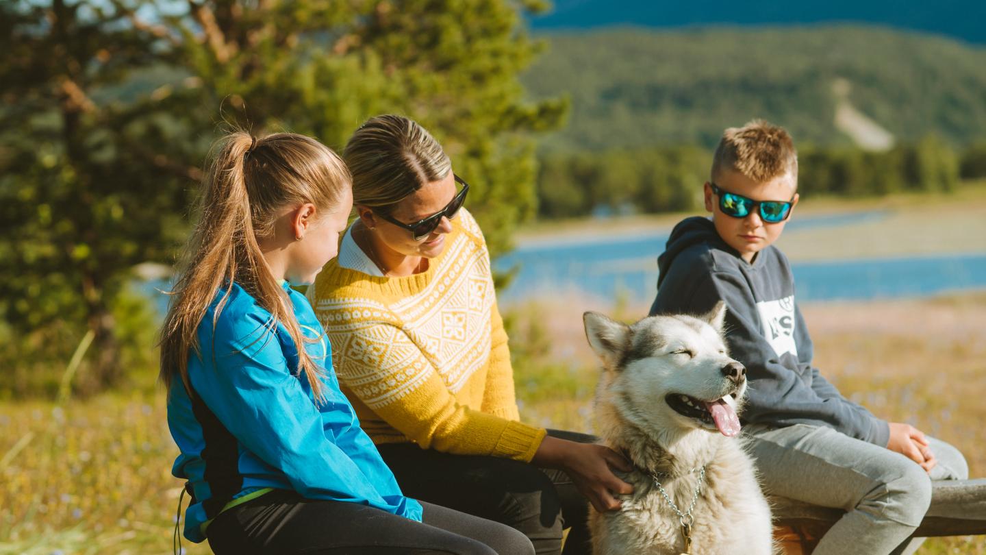 Mor og to barn sammen med hunden nyter sola ute på en benk i naturen