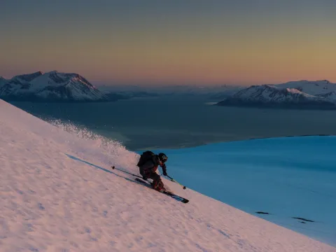 Mann kjører ned en fjellside på ski. I bakgrunnen kan du se havet og fjell.
