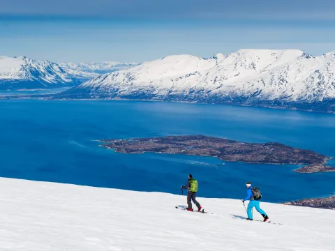 To personer går på ski. I bakgrunnen kan du se havet og fjell.