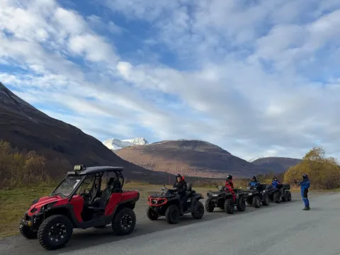 Mange firhjulingen på rekke og rad etter en grusvei. I bakgrunnen kan du se et jorde og høye fjell.