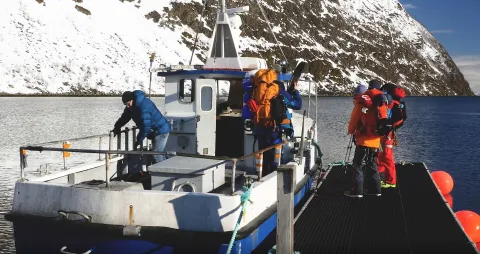 3 skifolk som holder på å gå ombord i en liten båt som ligger ved en flytebrygge. Kapteinen ombord står og gjør ting klart