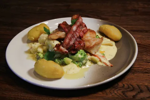 Hvit fisk med stekt bacon på og grønnsaker under. Tre poteter fordelt rundt på tallerkenen.