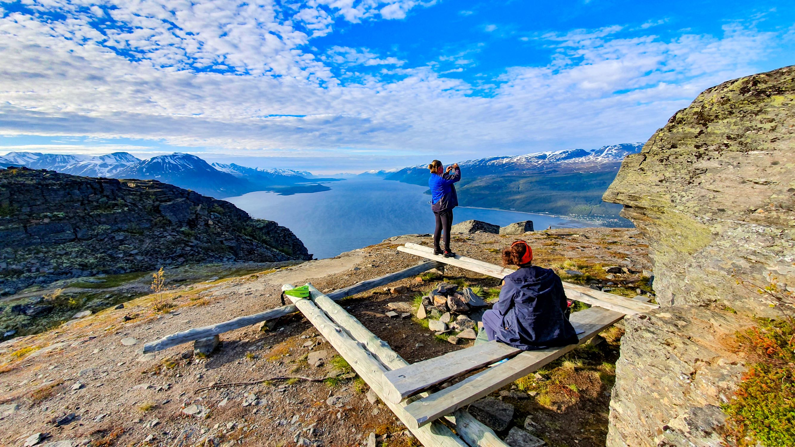 To personer ved en sittegruppe som nyter utsikten ut over lyngenfjorden, lyngsalpene i bakgrunnen